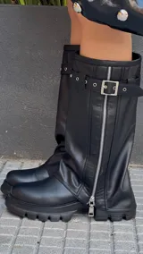 Botas de caña alta color negro, con suela track y cierre lateral con cremallera. Presentan una correa con hebilla alrededor de la caña.