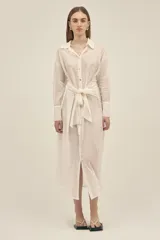 Vestido camisero largo color crema, de algodón, con cuello clásico, mangas largas con puños abotonados, cierre frontal con botones nacarados y lazo para atar en la cintura.