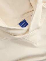 Canguro color crema con capucha, mangas largas y puños acanalados. Estampado frontal con texto "Originals Studio" en color marrón.