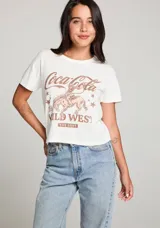 Remera color crema de corte cropped, con cuello redondo y mangas cortas. Estampado frontal con el logo de Coca-Cola, un jinete a caballo y la frase "The Wild West The Best" en color marrón.