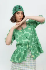 Camisa verde con estampado de flores, confeccionada en lino o popelina, con cuello redondo y mangas cortas abullonadas. Se cierra con tres lazos en el frente y tiene un corte con volantes en la parte inferior.