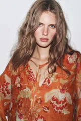 Camisa arrugada de ramio color naranja con estampado floral en tonos rosados y blancos. Tiene cuello redondo con escote pico ajustable con lazadas, mangas 3/4 y bajo asimétrico con aberturas laterales.