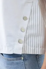 Remera blanca de corte holgado con cuello en V y mangas cortas. Presenta detalle de tela a rayas verticales en los laterales con botones decorativos.