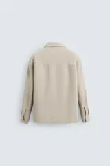 Sobrecamisa color beige de corte relaxed fit, con cuello solapa, manga larga con puños abotonados, bolsillos de plastrón con solapa en el pecho y bolsillos de vivo en la cadera. Cierre frontal con botones.