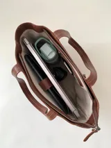 Cartera tipo tote de cuero graneado color negro, con textura uniforme y suave al tacto. Tiene manijas de 55 cm, bolsillo frontal con broche imantado, bolsillo trasero con cierre metálico, cierre principal metálico, forro interior y tres bolsillos internos (uno con cierre, uno plaqué y uno tipo sobre con solapa y botón).