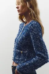 Chaqueta corta azul con tejido de punto entrelazado, cuello redondo, manga larga, bolsillos delanteros de plastrón y cierre frontal con botones dorados.