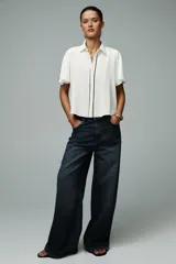 Camisa blanca de corte recto, con cuello solapa y mangas cortas. Presenta vivos negros a contraste en el frente y aberturas laterales en el ruedo. Cierre frontal con botones ocultos.