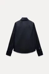 Camisa de denim azul oscuro con cuello con lazos, mangas largas con puños abotonados, bajo redondeado y cierre frontal con botones ocultos.