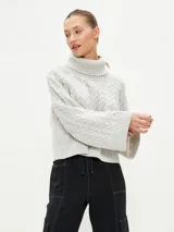 Polerón de tejido de punto color gris claro, con cuello alto y diseño de trenzas en el cuerpo y mangas. Corte holgado y puños anchos.