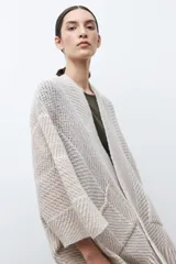 Ruana de tejido de punto en tonos beige y blanco, con patrón de espiga. Presenta cuello en V, mangas amplias, dos bolsillos frontales y flecos en el dobladillo.