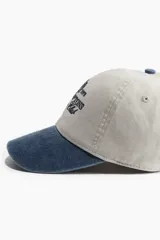 Gorra de béisbol color beige con visera azul denim, bordado frontal con el texto "Est. 1973 The Hamptons Country Club" y un escudo.