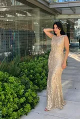 Vestido largo sin mangas, color dorado, con base beige y diseño de encaje o brocado con brillo. Corte ajustado al cuerpo que cae recto hasta el suelo.