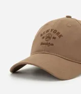 Gorra de sarga color beige con visera curva, bordado frontal con la inscripción "New York 1996 Brooklyn" en marrón. Ajuste trasero con hebilla metálica.