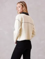 Chaqueta de corte boxy en color crema, confeccionada en tejido de punto suave con detalles en cuero sintético negro en cuello, solapas, cartera frontal y bolsillos. Presenta cierre frontal con botones metálicos plateados, hombros caídos y costuras decorativas horizontales.