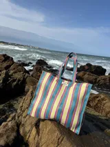 Bolso tipo tote grande con rayas verticales en celeste, blanco y bordó. Tiene doble asa corta en celeste con borde bordó, cierre superior y bolsillo interno.