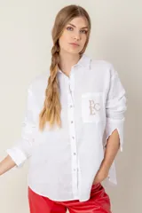 Camisa blanca de lino con corte holgado, cuello clásico, manga larga con puños abotonados, cierre frontal con botones y bolsillo en el pecho con bordado de monograma en color beige.
