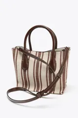 Bolso estilo bandolera con estampado a rayas verticales color marrón y crudo. Tiene asa de mano y asa fina extraíble con detalles metálicos. El bolso interior es a contraste de materiales con cierre mediante cremallera metálica. Cierre imantado.