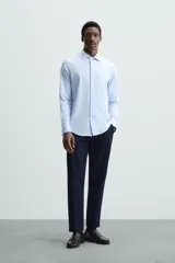 Camisa celeste a rayas verticales blancas, de corte recto, con cuello italiano abierto, mangas largas con puños abotonados y cierre frontal con botones.