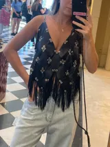 Top negro de cuero genuino, confeccionado a mano con técnica zero waste. Presenta un diseño con paneles unidos por ojales metálicos plateados y flecos largos en el ruedo inferior. Tiene tirantes finos ajustables.