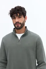 Buzo de algodón color verde musgo, con cuello alto y cierre frontal parcial. Presenta puños y cintura acanalados, y un pequeño logo bordado en el pecho.