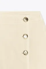 Falda pantalón corta color crudo, de tiro alto con delantero cruzado y cierre con cremallera oculta y botones.