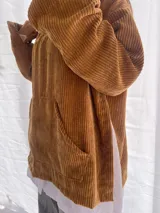 Buzo de pana fina color verde oliva, con diseño de corte holgado y mangas largas. Presenta una textura de cordón vertical característica de la pana y un pequeño parche decorativo en la parte superior de la espalda.