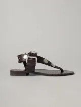 Sandalias planas de cuero color marrón, con tira entre los dedos, pulsera al tobillo con hebilla metálica y apliques de tachas circulares plateadas.