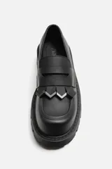Mocasín negro de cuero con antifaz y flecos con puntas metálicas en el empeine, horma cuadrada y suela con volumen.