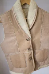 Chaleco de cuero color beige con interior de piel sintética color crema, cierre frontal con botones y bolsillos delanteros.