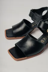 Sandalias negras de cuero con diseño minimalista y taco ancho geométrico en tono madera. La capellada lisa aporta una estética depurada y moderna, mientras que la pulsera con hebilla metálica ajustable asegura un calce firme y confortable.