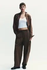 Camisa oversize marrón con rayas verticales blancas, cuello solapa, manga larga con puños y hombros caídos. Tiene un bolsillo de plastrón en el delantero, pinzas en la espalda y cierre frontal con botones.