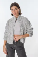Cardigan tejido de punto grueso color gris claro con diseño de trenzas, cuello alto y cierre frontal con cremallera.