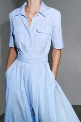 Vestido camisero midi de popelín a rayas verticales celestes y blancas, con cuello solapa, manga corta, bolsillos de plastrón con solapa en el pecho, cinturón de lazo ajustable y cierre frontal con botones.