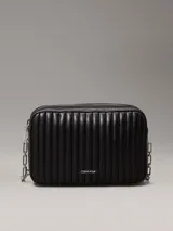 Cartera bandolera Calvin Klein negra con diseño acolchado de líneas verticales, correa de cadena plateada, cierre superior con cremallera y logo metálico frontal.