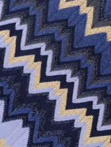 Chal tipo bufanda de punto con patrón de zigzag en tonos azules, lavanda y beige/dorado. Presenta un diseño multicolor con hilos brillantes.