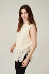 Chaleco de punto sin mangas color beige, con cuello alto tipo polera. Presenta un diseño de tejido con ochos y rombos, y terminación de flecos largos en el ruedo.