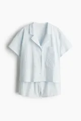 Pijama corto de dos piezas color celeste, en tejido liviano de algodón con broderie. Camisa de manga corta con cuello abierto, botones adelante, bolsillo superior abierto y bajo recto con aberturas laterales. Short con cintura elástica.