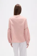 Blusa holgada de algodón color rosa pálido, con escote redondo y lazo ajustable en el centro. Presenta detalles de encaje calado alrededor del escote y en los puños de las mangas largas abullonadas.