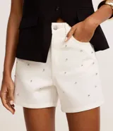 Short de cintura alta en sarga color blanco, con trabillas en la cintura, cierre tradicional, bolsillos, lunares de metal y strass aplicados.
