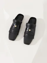 Mocasín tipo mule de cuero vacuno negro, con flecos y aplique metálico dorado en el empeine.