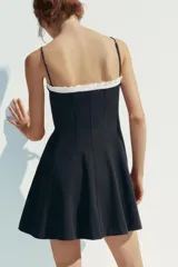 Vestido corto negro con tirantes finos ajustables, escote recto con volantes blancos y lazo decorativo. Cierre lateral con cremallera oculta.