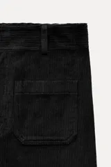 Pantalón ancho de pana color negro, de tiro medio, con trabillas en la cintura, bolsillos delanteros y cierre frontal con cremallera, botón y gancho metálico.
