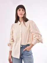 Camisa color crema con rayas verticales, cuello clásico, mangas largas abullonadas con puños elásticos y cierre frontal con botones. Corte holgado.