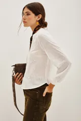 Camisa blanca de manga larga con cuello mao y escote en V. Presenta bordados florales en blanco y dorado en el frente.