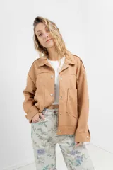 Chaqueta color beige de corte recto, estilo camisa, confeccionada en gamuza con elastano. Presenta cuello camisero, cierre frontal con botones metálicos, bolsillos grandes en el frente y charreteras en los hombros.