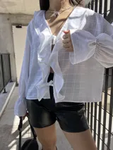 Camisa blanca traslúcida con diseño a cuadros, escote en V con lazo, mangas largas abullonadas con puños elásticos y corte holgado.