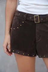 Short de gamuza color marrón oscuro, con tiro alto, cinturón incorporado con hebilla metálica y tachas doradas decorativas en los bordes de los bolsillos y el ruedo.