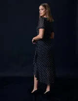 Vestido midi de gasa color negro con lunares blancos, con mangas cortas, escote redondo y ruedo asimétrico. Presenta un frunce lateral con lazo de ajuste.
