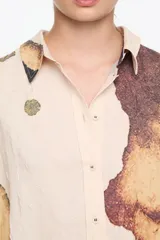Camisa de corte recto con cuello camisero y botones frontales. La tela presenta un efecto arrugado o texturizado en tono beige claro, con grandes bloques de estampado abstracto en tonos tierra (marrón, ocre, verde oliva) que simulan manchas o texturas orgánicas. Las mangas son largas y una de ellas tiene un detalle en negro liso en el puño.