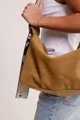 Bolso de hombro tipo hobo de gamuza color marrón óxido, con correa ancha ajustable de color negro y herrajes metálicos plateados. El nombre de la marca "GANESHA" está grabado sutilmente en el cuerpo del bolso.